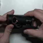 一雙手正握持並調整一台黑色 Mio 品牌行車記錄器,機器鏡頭周圍有紅色圓圈設計,底部裝有吸盤支架。背景為簡約的白色桌面與一盆綠色小植物