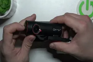 一雙手正握持並調整一台黑色 Mio 品牌行車記錄器,機器鏡頭周圍有紅色圓圈設計,底部裝有吸盤支架。背景為簡約的白色桌面與一盆綠色小植物